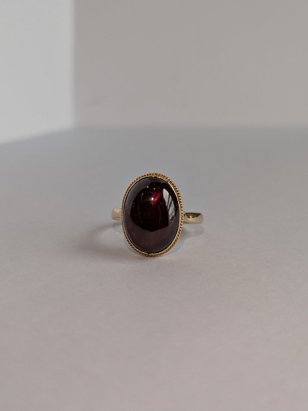 Front on Shot of yellow gold ring set with deep red oval cabochon cut garnet bezel set, ring laying flat on white background