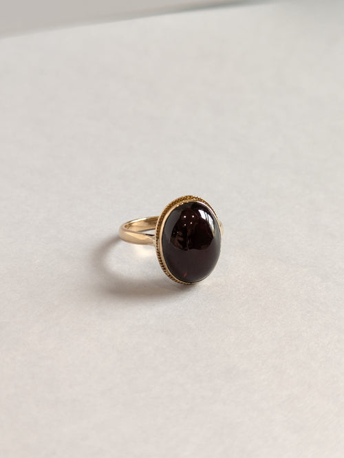 Slight Off Angle Front on Shot of yellow gold ring set with deep red oval cabochon cut garnet bezel set, ring laying flat on white background
