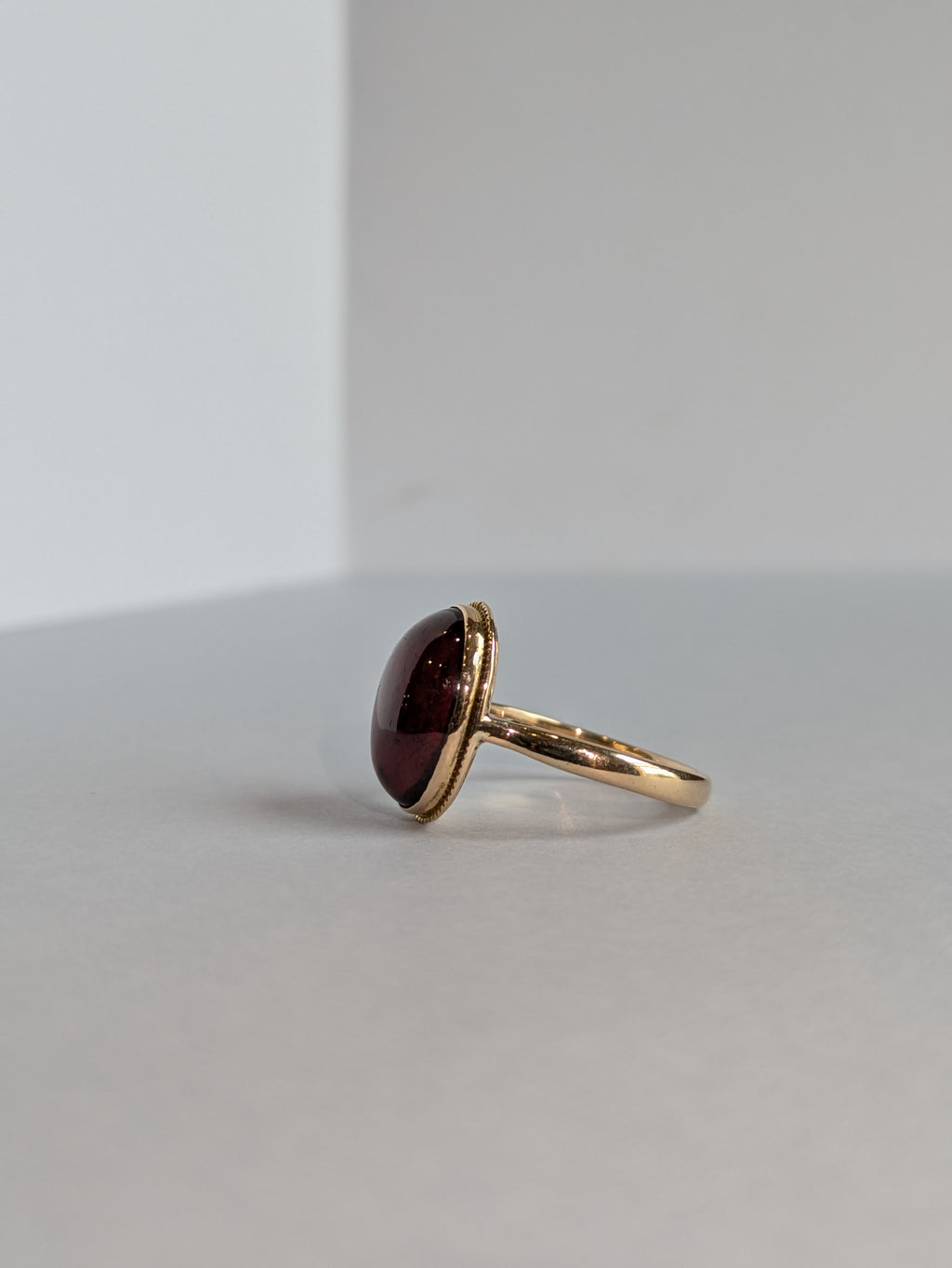 Side View Camera Shot of yellow gold ring set with deep red oval cabochon cut garnet bezel set, ring laying flat on white background 90 degrees away from camera