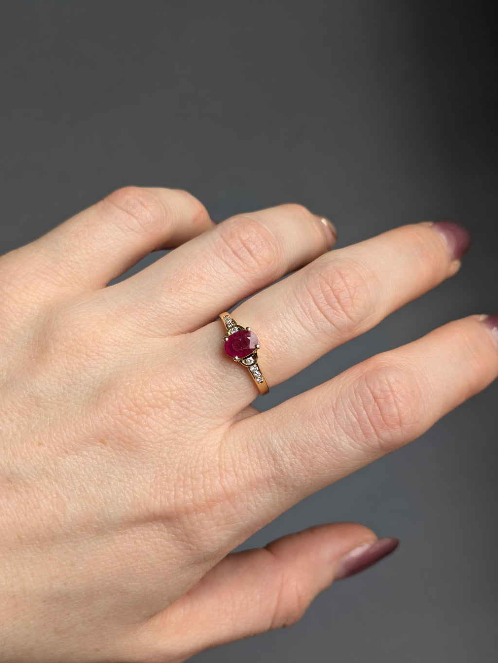 Fair-skinned Hand wearing a Yellow Gold Ring set with Deep Red Oval Ruby and Round Diamonds on the shoulders.