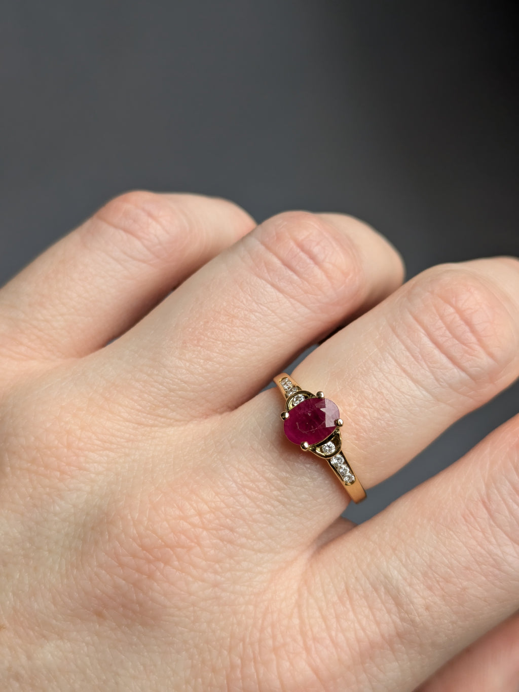 Closeup of Fair-skinned White Hand wearing Deep Red Oval Ruby Claw Set in Yellow Gold Ring with Round Brilliant Cut Diamonds Set in Shoulders