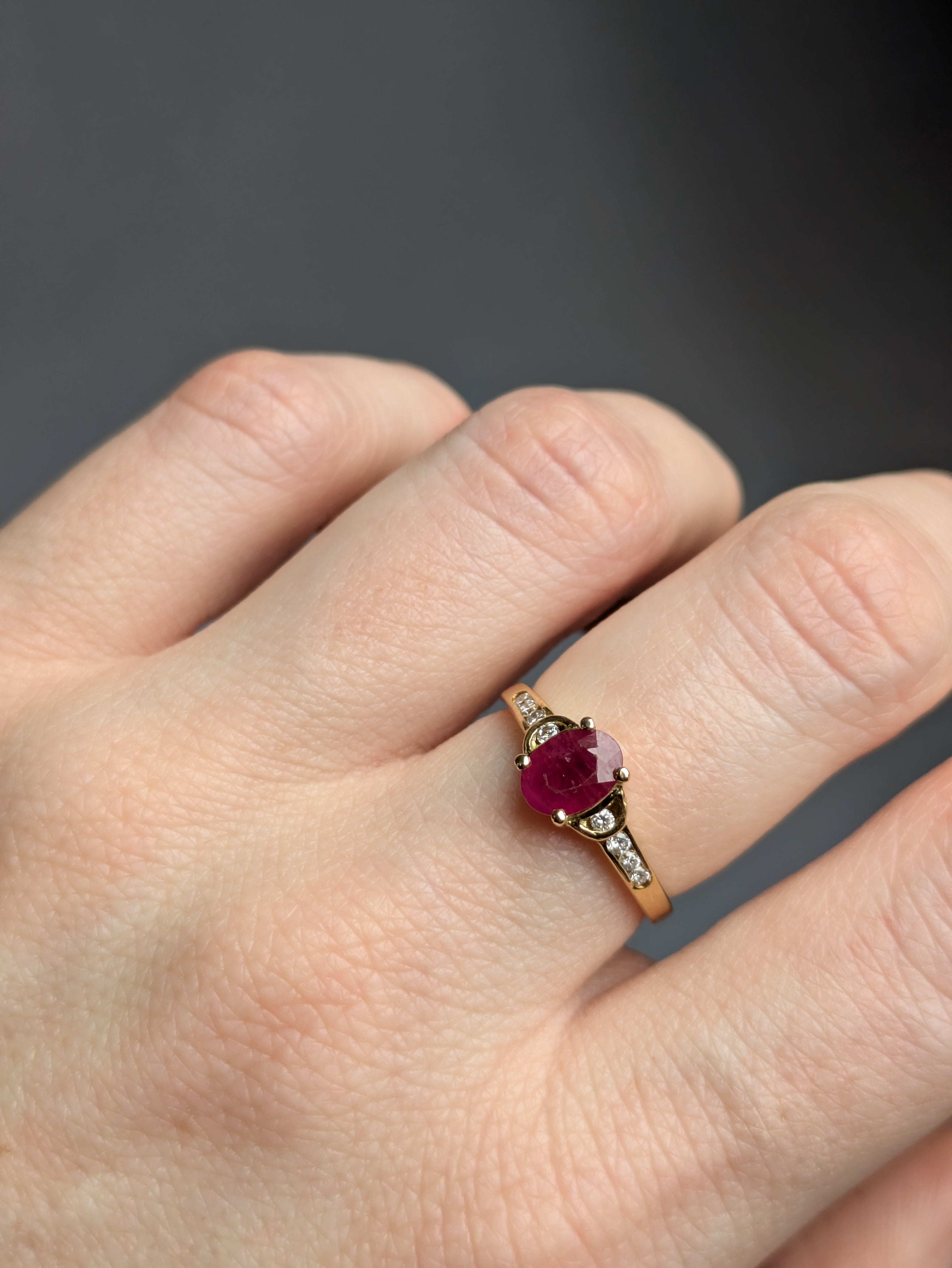 Closeup of Fair-skinned White Hand wearing Deep Red Oval Ruby Claw Set in Yellow Gold Ring with Round Brilliant Cut Diamonds Set in Shoulders