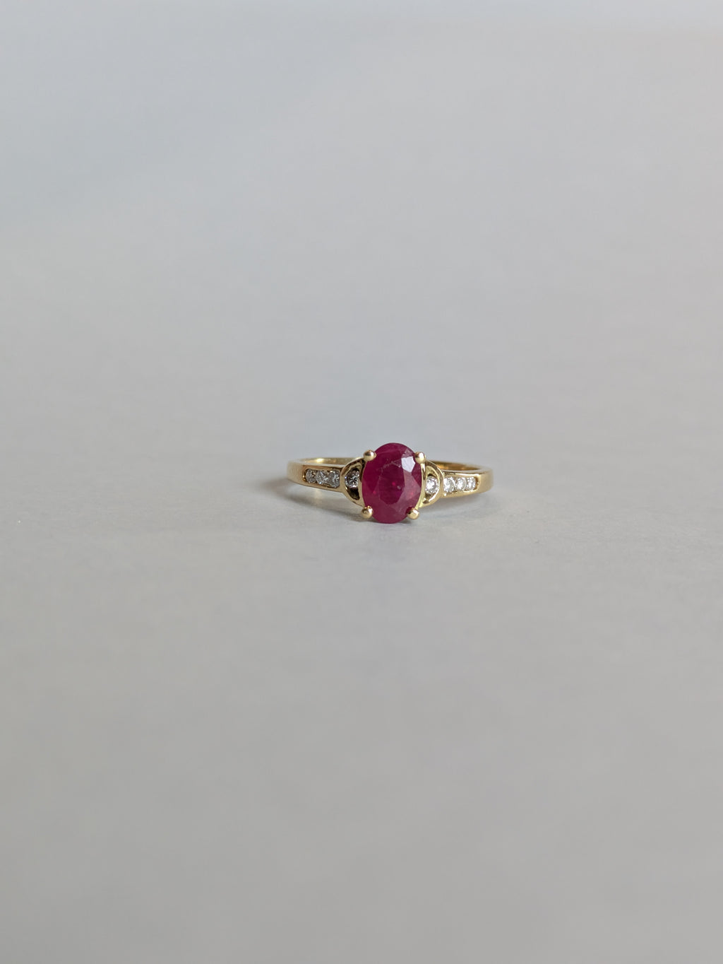 Deep Red Oval Ruby Claw Set in Yellow Gold Ring with Round Brilliant Cut Diamonds Set in Shoulders Laying Flat on White Background