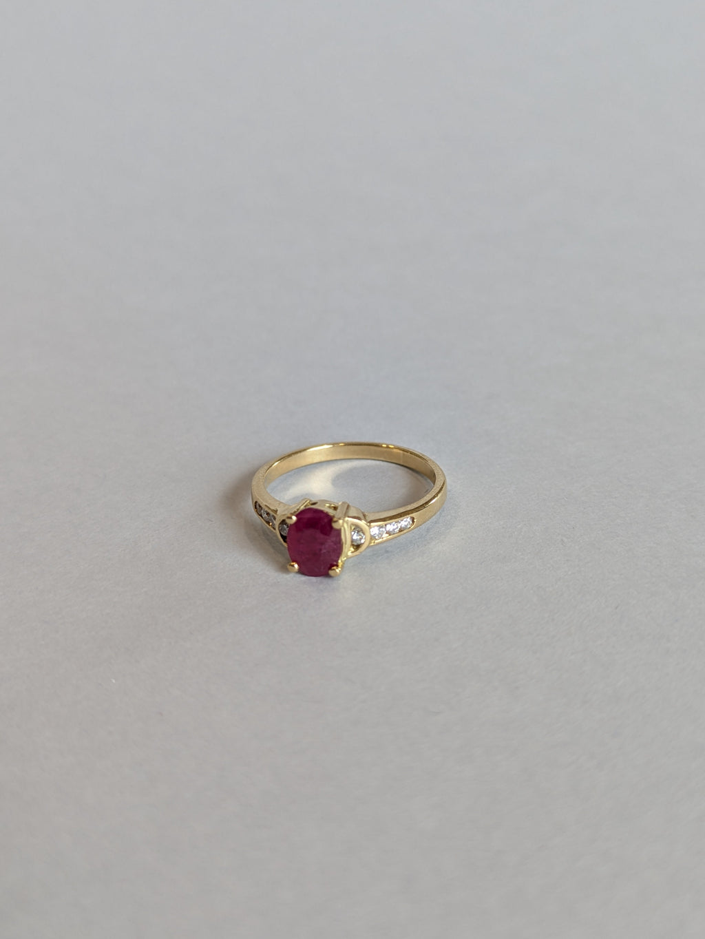 Deep Red Oval Ruby Claw Set in Yellow Gold Ring with Round Brilliant Cut Diamonds Set in Shoulders Laying Flat on White Background Facing Away from Camera