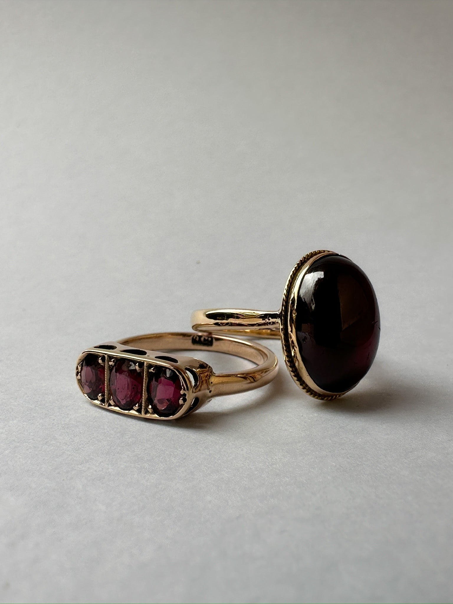 Yellow gold garnet rings, one with three stones, one with a large cabochon

Two subjects camera shot with yellow gold ring set with deep red oval cabochon cut garnet bezel set, ring laying flat on white background